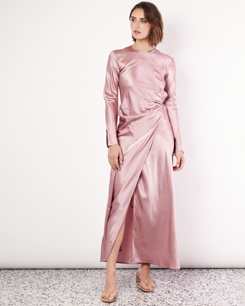 The Long Sleeve Wrap Dress boasts a fluid form and elevated style, featuring gathered detailing and a leg split. It is crafted from 100% Crushed Acetate in a Blush hue. By Romy, now available at After Eight. 