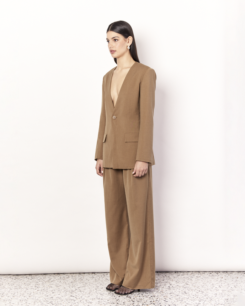 The Collarless Blazer is a versatile tailored silhouette featuring two front pockets, a button closure and a collarless neckline. It is fully lined and is crafted from a soft wool blend in Tan. By Romy, now available at After Eight. 