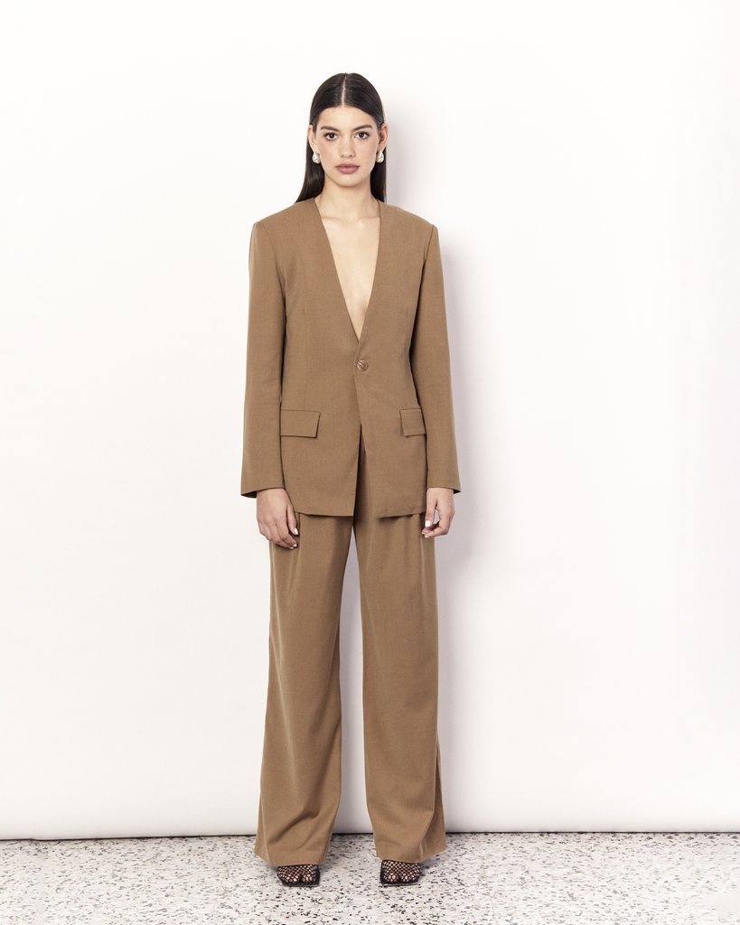The Collarless Blazer is a versatile tailored silhouette featuring two front pockets, a button closure and a collarless neckline. It is fully lined and is crafted from a soft wool blend in Tan. By Romy, now available at After Eight. 