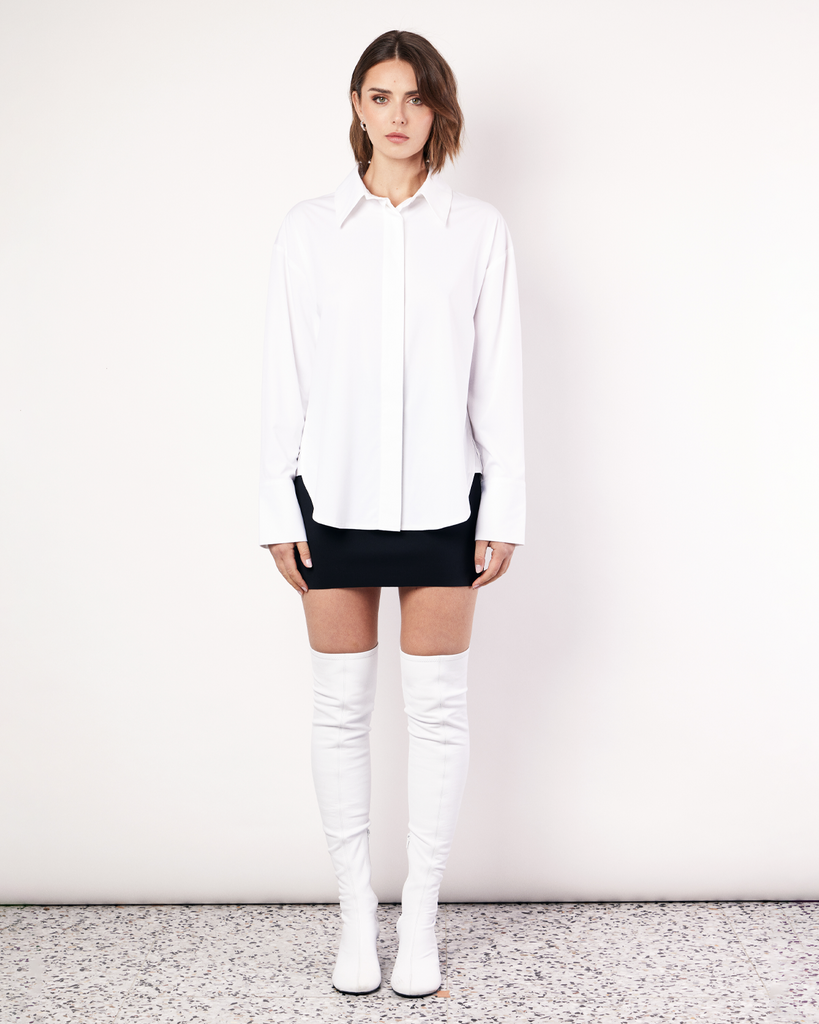 The Classic Shirt in white is a wardrobe icon. Cut oversized with a scooped hem and elongated cuff, this shirt is the perfect pairing for achieving a timeless look. Crafted from a regenerated cotton blend with a touch of stretch, it's not only stylish but also less likely to crease compared to 100% cotton, ensuring you stay effortlessly chic all day long. By Romy, now available at After Eight. 