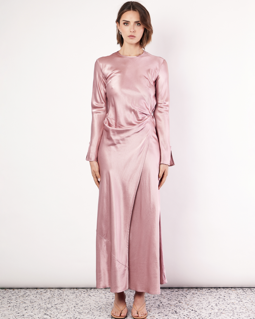 The Long Sleeve Wrap Dress boasts a fluid form and elevated style, featuring gathered detailing and a leg split. It is crafted from 100% Crushed Acetate in a Blush hue. By Romy, now available at After Eight. 