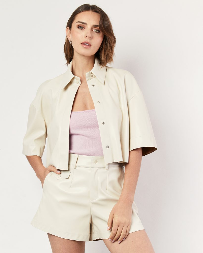 The Vegan Leather Cropped Shirt in cream is meticulously crafted from supple vegan leather, offering a luxurious feel against your skin. It's expertly tailored for an oversized silhouette, ensuring both comfort and style. Style with the coordinating Vegan Leather Shorts. By Romy, now available at After Eight. 