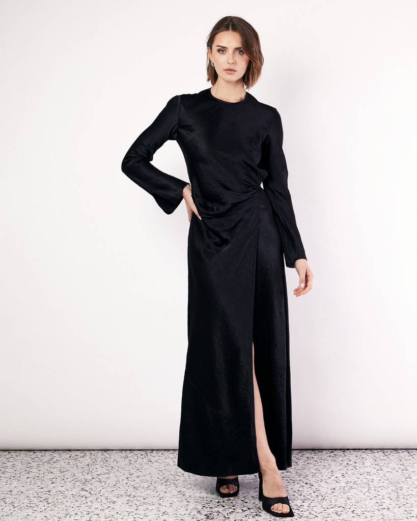 The Long Sleeve Wrap Dress boasts a fluid form and elevated style, featuring gathered detailing and a leg split. It is crafted from a 100% Crushed Acetate in Black. By Romy, now available at After Eight. 