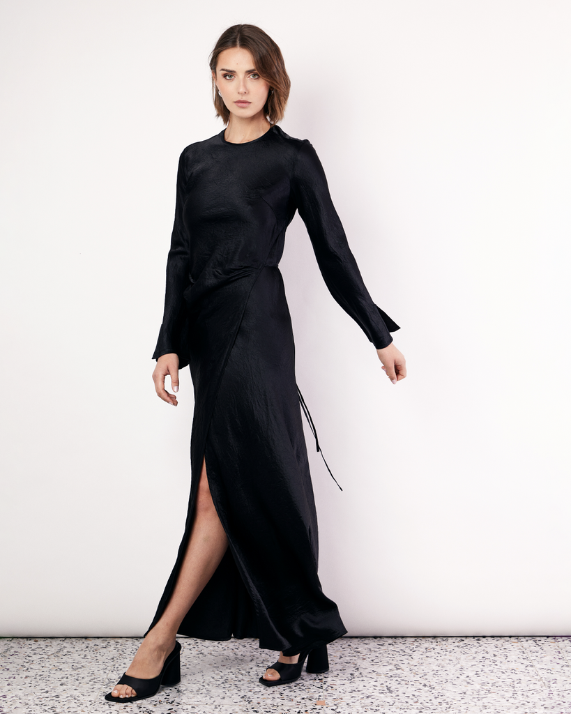 The Long Sleeve Wrap Dress boasts a fluid form and elevated style, featuring gathered detailing and a leg split. It is crafted from a 100% Crushed Acetate in Black. By Romy, now available at After Eight. 